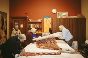 A classroom with book shelves on the wall and tall cupboards.  At a large table covered with a white cloth two ladies roll a patterned red, black and white textile around a tissue paper covered roll.  A third lady records the work on a piece of paper and a four watches on from the back.