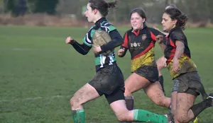 A mud splattered female student in green, black and white holding a rugby ball runs away from two opponents