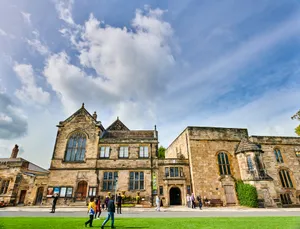 A series of historic stone buildings of different shapes and sizes sit behind a roadway. There is a large lawn in the front.