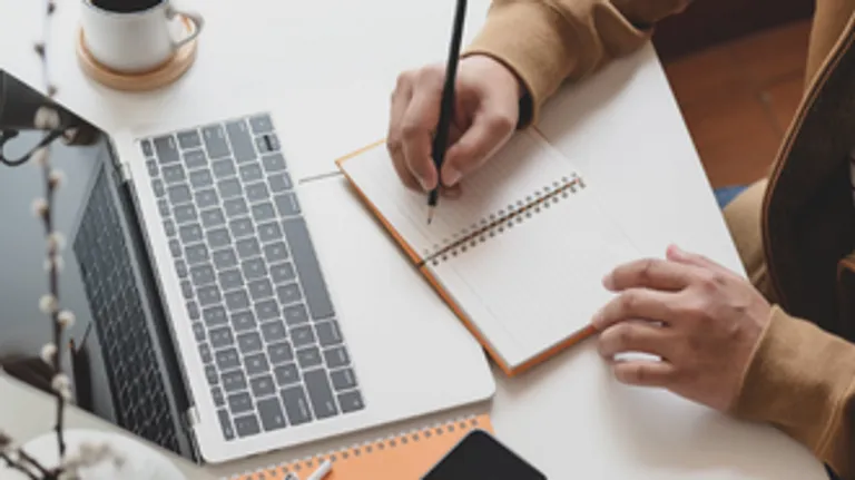 Close up of a persons hands taking notes in a notebook in front of their laptop