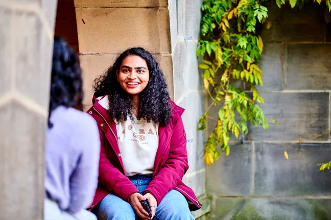 Two students sitting in the courtyard of St Marys on a sunny day having a friendly conversation while sitting and leaning against limestone buildings