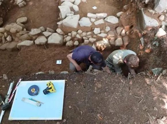 Two people crouched in an archaeological trench, drawing a section of deposits. Behind them are the remains of a building. On the edge of the trench is some drawing equipment.