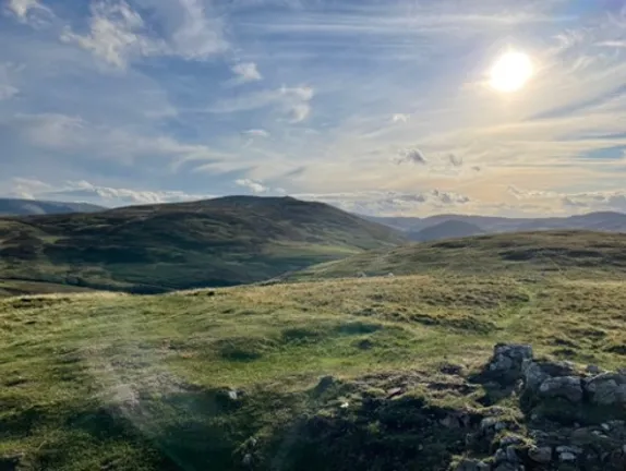 The sun is high over grassy hills. The sky is blue with cirrus clouds. In the foreground are some rocks.