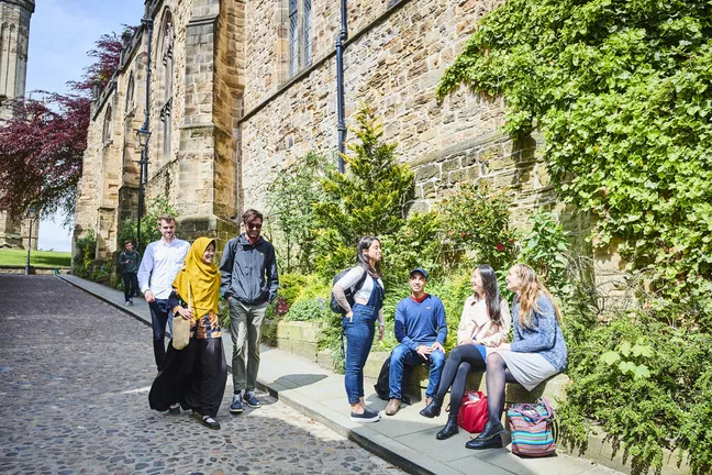 A group of students are talking in the sunshine, in front of historical buildings in Durham city.