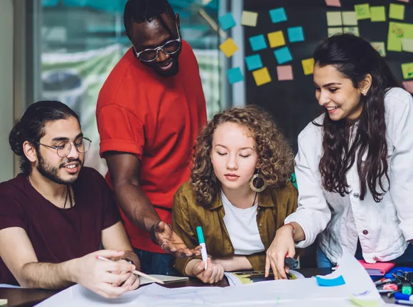Fours adults, 2 men, 2 women, around desk with pens and paper.