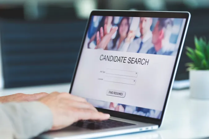 A hand on the keyboard of an open laptop. On the screen, it reads, 'Candidate Search'.