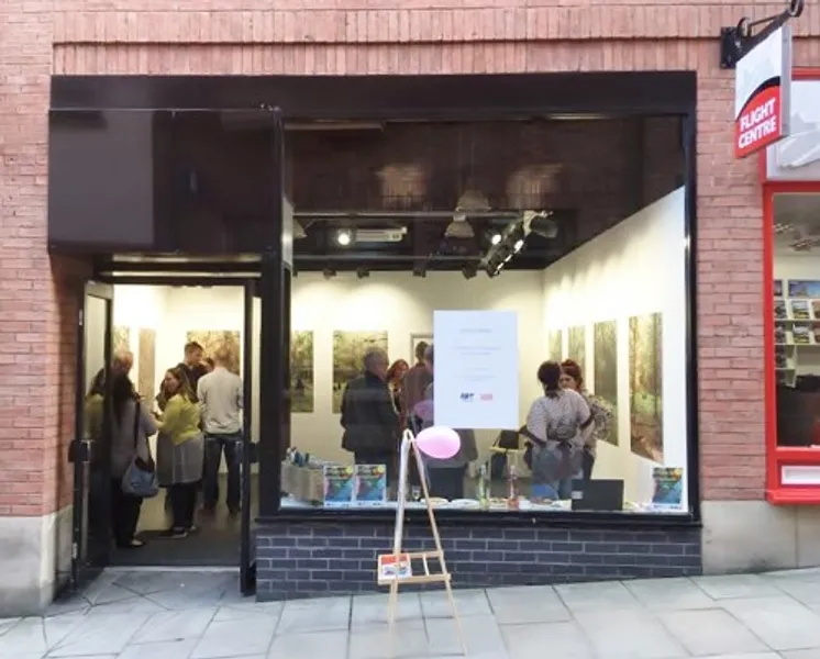 Looking into a large shop window from outside, inside the shop is an art exhibition on the walls, groups of people stand inside the shop chatting. Outside the shop is an A-board in side profile with a pink balloon attached to it.