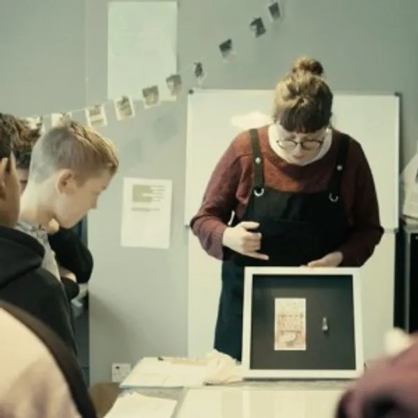 Photograph of an art display class – a member of the Learning and Engagement Team is holding up an artwork and pointing to it while talking about to a group of students who are gathered around it.