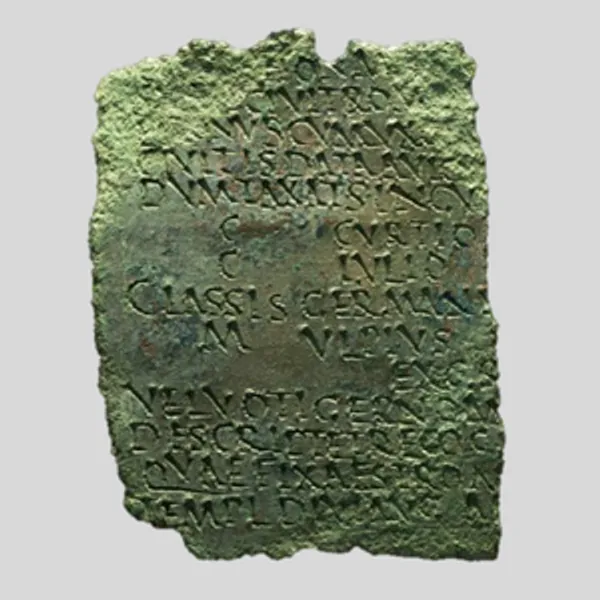 Photograph of a fragment of the Lanchester Diploma – a flat piece of corroded and damaged green metal covered in a Latin inscription that has been punched in and written in block capitals.