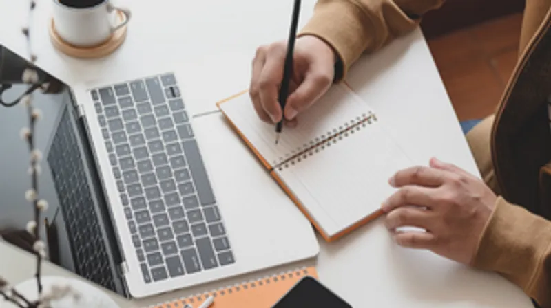 Close up of a persons hands taking notes in a notebook in front of their laptop