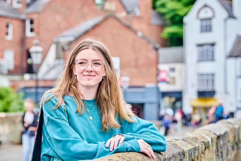 A young woman with long blonde hair and glasses wearing a blue jumper with her hands over the side of a bridge