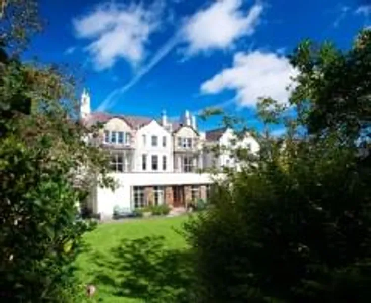 Image shows exterior of St Cuthberts Society from the garden.  It is a lovely day with a blue sky..  Trees frame the white building.