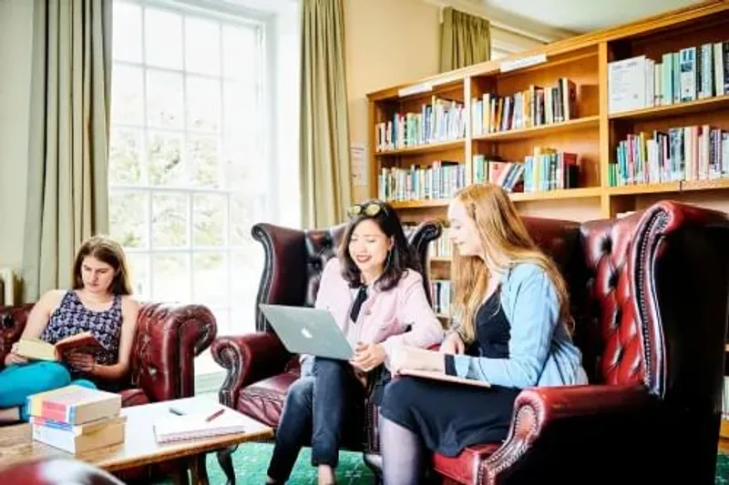 A group of students study using books and a laptop on a comfy sofa in St Mary's Library, behind is a bookcase full of books.