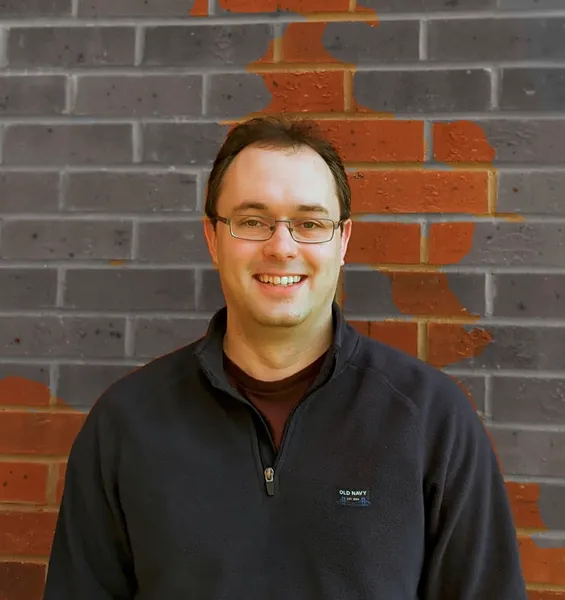 A portrait shot of Dr Doupe. Dr Doupe is wearing a dark top, standing against a grey brick wall, and smiling at the camera.