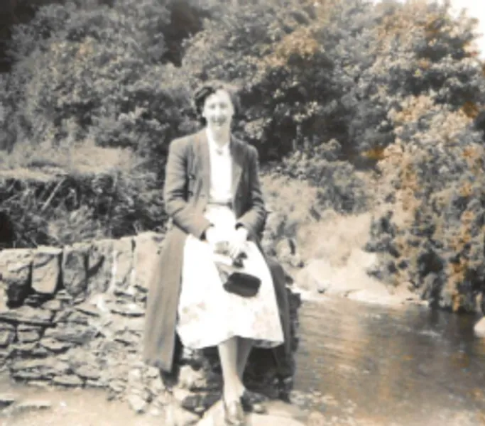 A black and white photo of alumna Margaret Bloomfield sitting on a rock by a river.