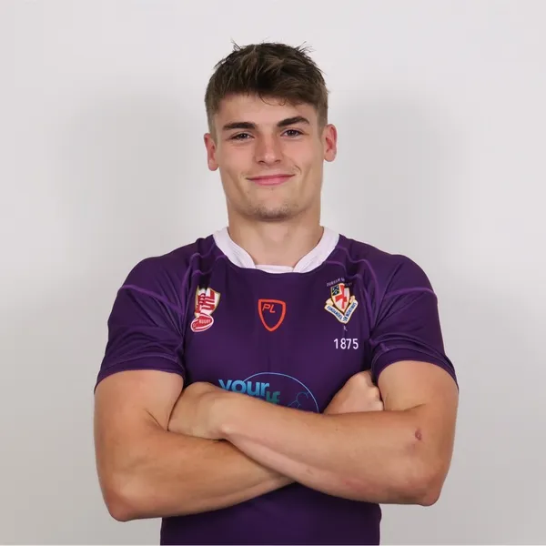 A photo of a man wearing a purple rugby top smiling straight into the camera with folded arms