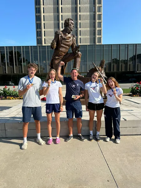 A mixed group of men and women standing in front of a statue in the sunshine wearing or holding gold medals while celebrating