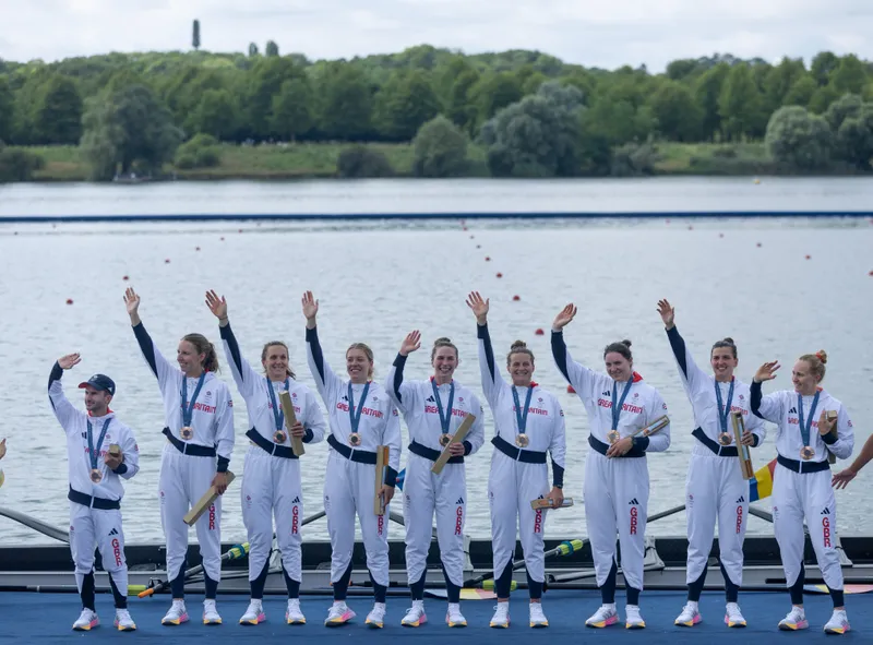 A group of female rowers and their cox in white tracksuits jumping in the air with their hands up