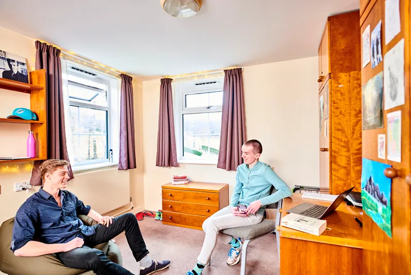 two students sitting in a bedroom, one in a chair and one on a beanbag, having a friendly conversation while relaxing