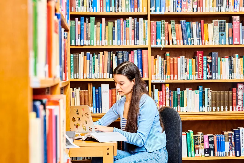 Student looking at book in front of bookshelf