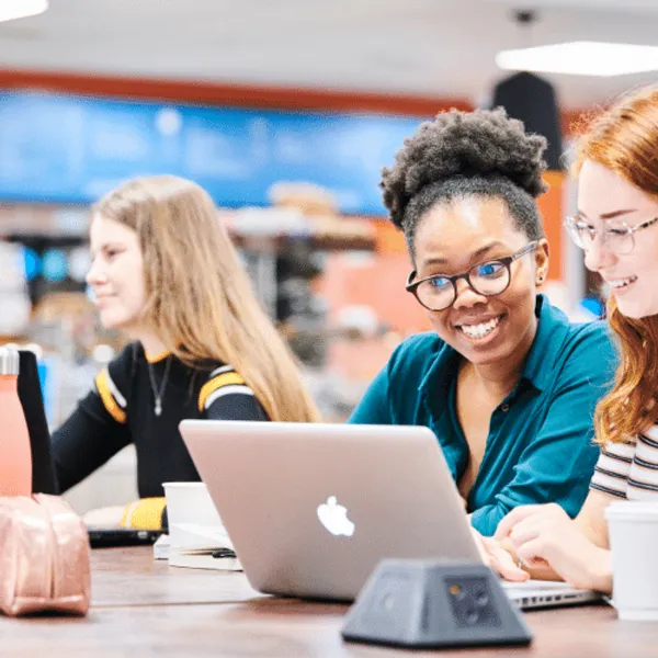 Two students looking at a laptop with a student in the background
