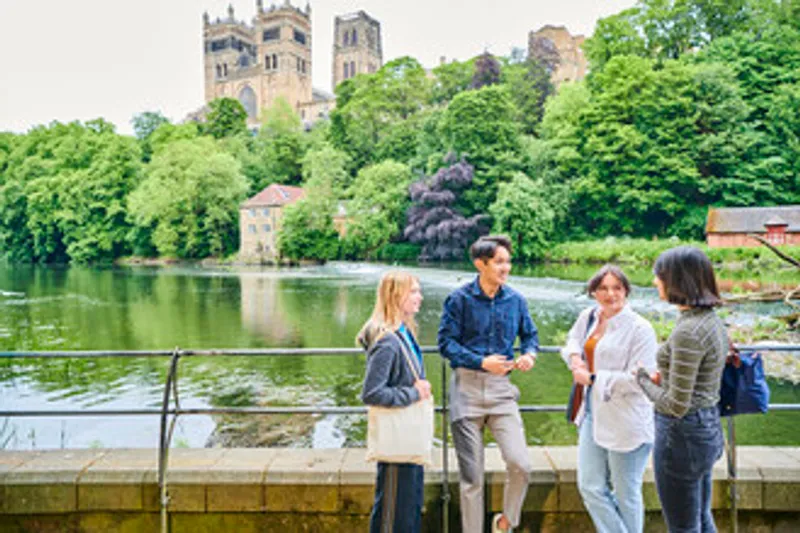 Students lean on some railings as they chat - the River Wear is behind them and an old red brick building (Old Fulling Mill) and the Cathedral can be seen on the other side. Lots of green trees in full leaf plus one dark purple copper beech tree.