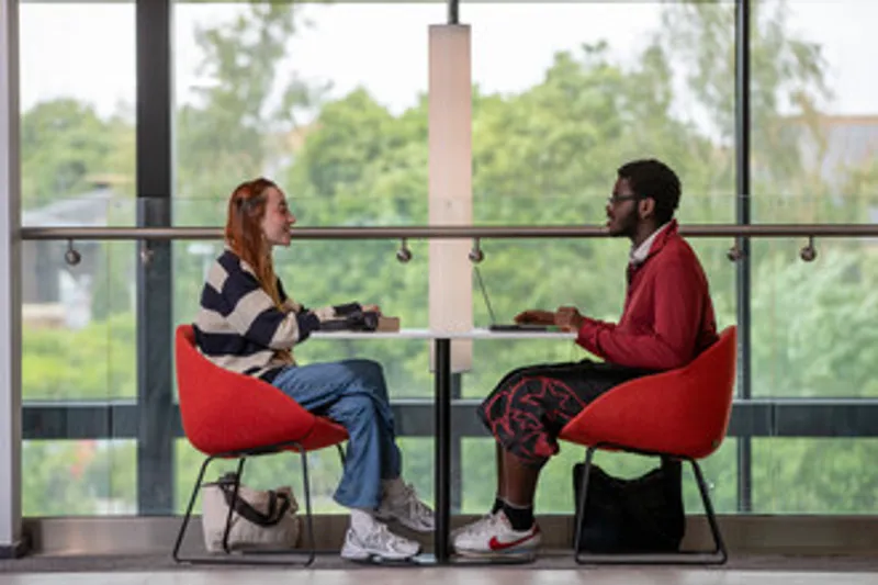 Two students sit opposite each other at a table. They are inside a modern building with lots of large windows