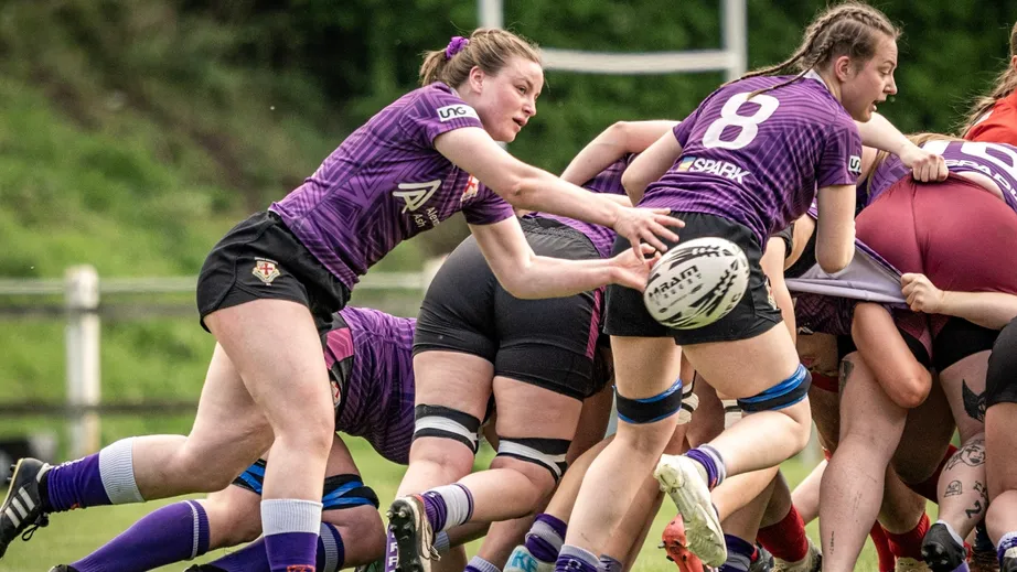 Two teams of female rugby players wearing purple tops, and red and white tops, playing against each other