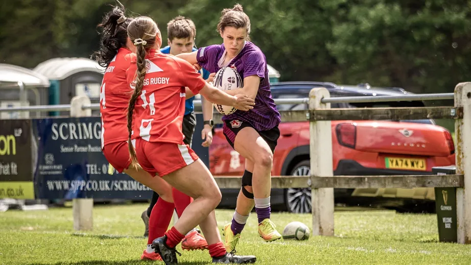 Two teams of female rugby players wearing purple tops, and red and white tops, playing against each other