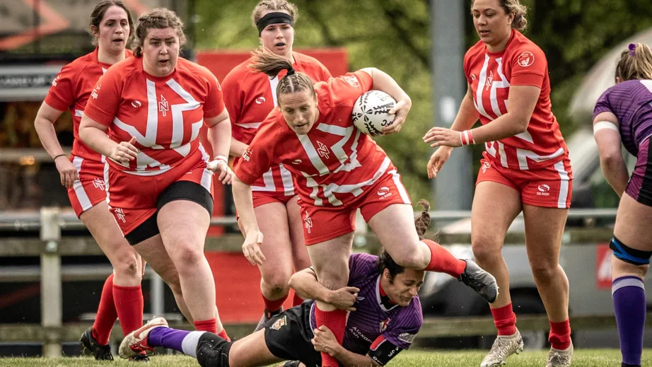 Two teams of female rugby players wearing purple tops, and red and white tops, playing against each other