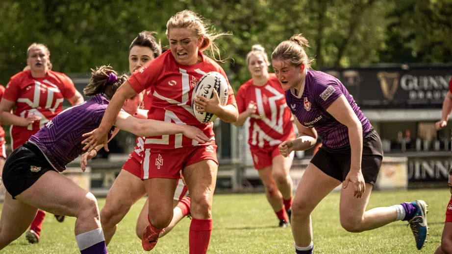 Two teams of female rugby players wearing purple tops, and red and white tops, playing against each other