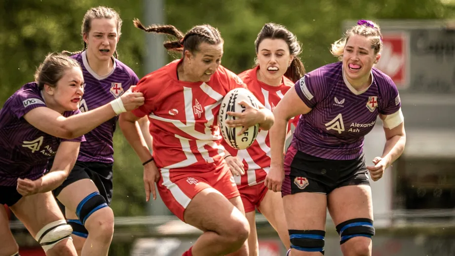 Two teams of female rugby players wearing purple tops, and red and white tops, playing against each other