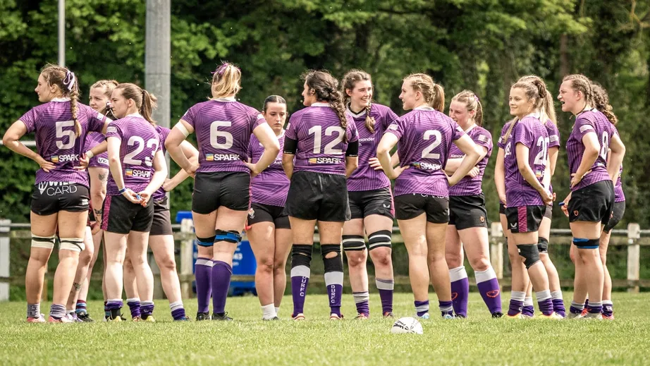 A team of female rugby players wearing purple tops, standing in a group with ttheir backs to the camera