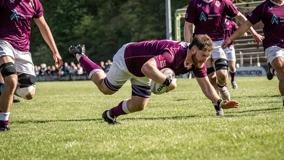A rugby player wearing a purple top and holding a rugby ball diving towards the ground.