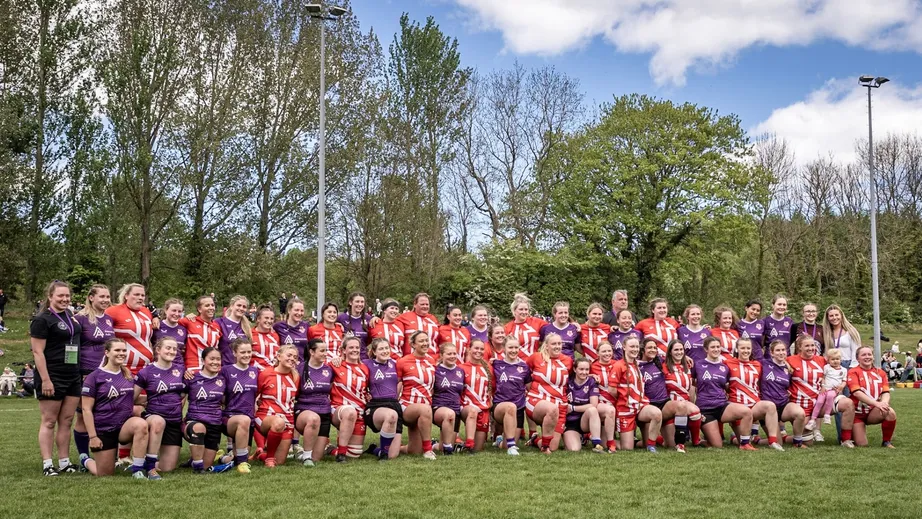 Two teams of female rugby players wearing purple tops, and red and white tops standing in a long line for the camera.
