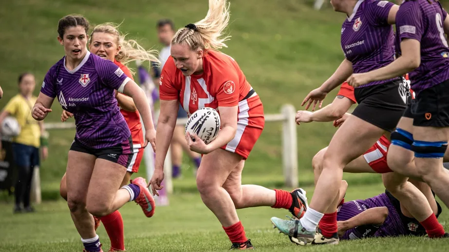 Two teams of female rugby players wearing purple tops, and red and white tops, playing against each other