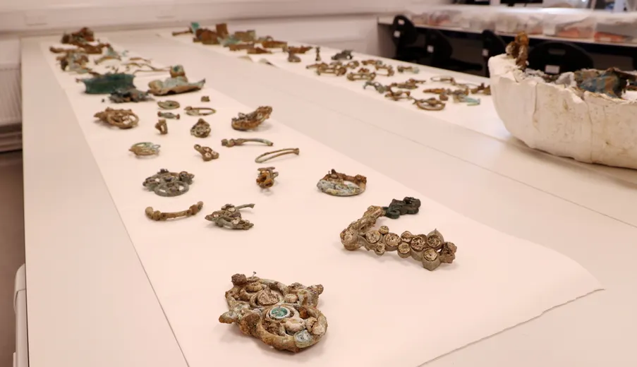 A variety of artefacts from the Melsonby Hoard laid out on a white table, including various pieces of corroded metalwork and a damaged cauldron.