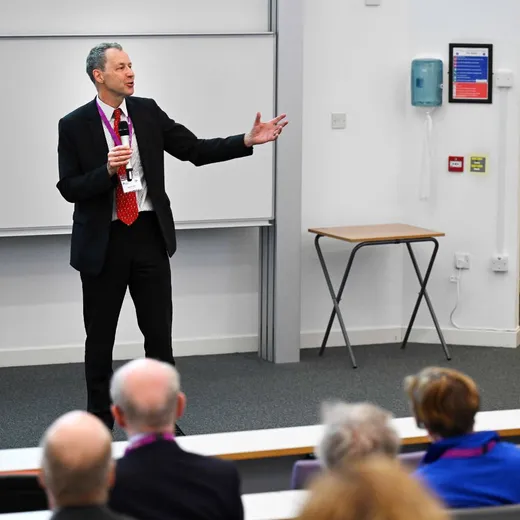 A man speaks with a microphone, with backs of heads of the audience in the foreground