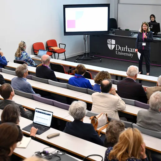 A woman speaks with a microphone, with backs of heads of the audience in the foreground, in tiered lecture hall seating