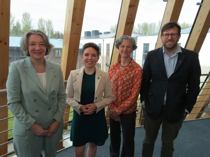 Four people looking at the camera, smiling. Behind them is a glass wall, through which can be seen other buildings and a grassed area