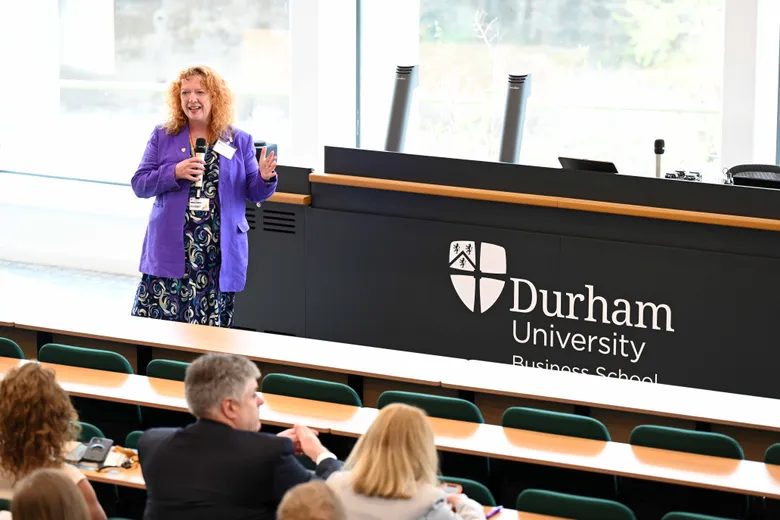 Woman in front of presentation desk, speaking to audience in tiered seating, backs of heads visible