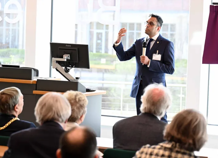 Man holding microphone, speaking to audience in tiered seating, backs of heads visible