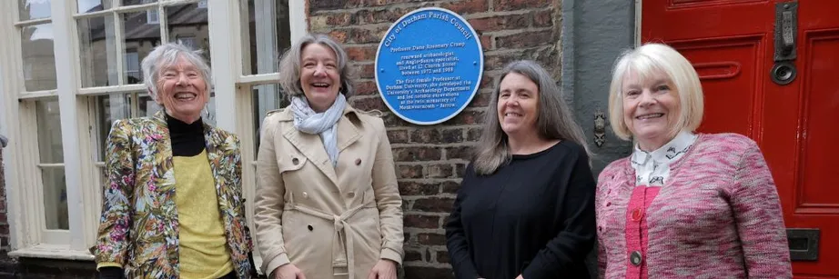 A group of women smile at the camera as they stand in front of a circular metal blue plaque fixed to a red brick wall.