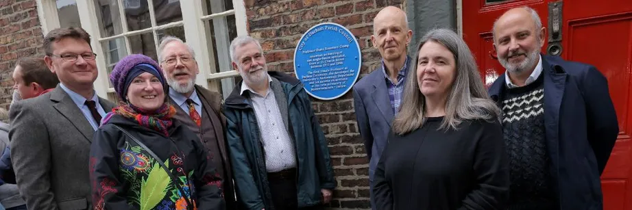 A group of men and women stand in front of a circular blue metal plaque set against a red brick wall.